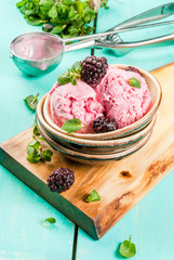 Berry ice cream in a small ice-cream bowls with blackberries and mint on a table, with berries and mint leaves and spoon on blue wooden table.