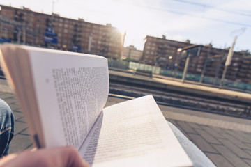 Young adult commuter seated on the railroad station platform, holds a book waiting for the train.