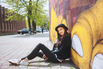 stylish young woman in hat sitting near the wall with graffiti