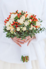 wedding bouquet with white lace ribbon, white roses and peonies, red berries and greens of eucalyptus in the hands of the bride, close-up