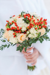 wedding bouquet with white lace ribbon, white roses and peonies, red berries and greens of eucalyptus in the hands of the bride, close-up