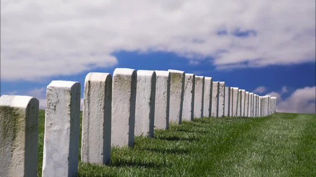 White military tombstones in a line across a grassy hill with time lapse motion clouds against a deep blue sky 