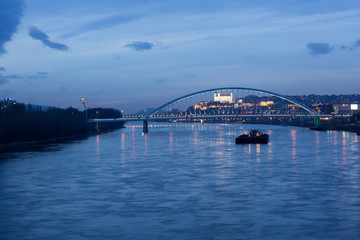 Naklejka premium Old town, riverside, Bratislava castle, Apollo Bridge, UFO Bridge, New Bridge with cargo ship in foreground shoot during dusk from above river Danube, Slovakia