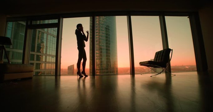 young woman businessman ceo talking on the phone, negotiating the panoramic windows in office skyscraper, sunset, dolly shot