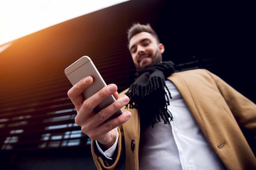 Handsome young man in coat and scarf texting friend with wide smile on face.