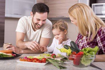 Young couple with kid having fun in kitchen while preparing fresh vegetable salad