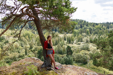The girl in the red sweater and shorts standing at the cliff leaning on a tree and enjoy the landscape and feel the freedom