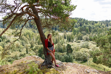 Naklejka na ściany i meble The girl in the red sweater and shorts standing at the cliff leaning on a tree and enjoy the landscape and feel the freedom. Looking at the camera and smiling.