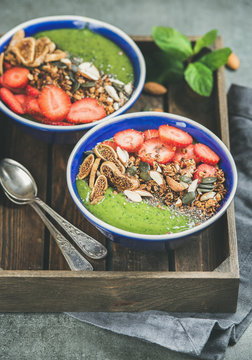 Healthy breakfast. Green smoothie bowls with strawberries, granola, chia and pumpkin seeds, dried fruit and nut in wooden tray over grey concrete background, selective focus. Vegan, raw food concept
