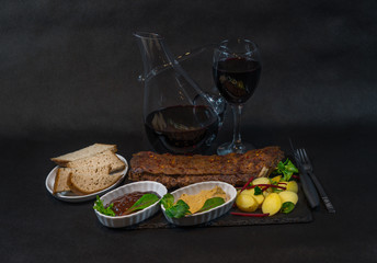 rack of pork ribs with potatoes, sauces, bread and red wine in a carafe and glass