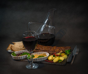 red wine in the glass on the left, rack of pork ribs with potatoes, sauces, bread and red wine in a...