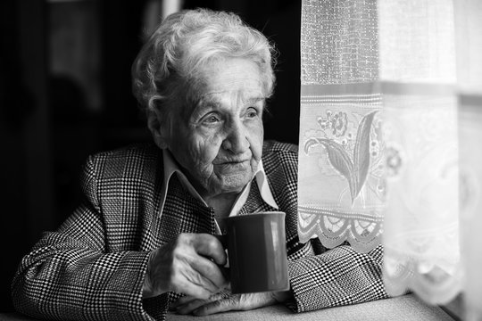 Elderly woman drinking tea near the window. Black-and-white photo.