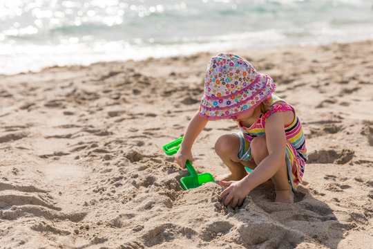 Little girl in a hat and sundress plays with a toy shovel on the beach