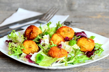 Fried mashed potato balls on a plate, fork, knife, napkin on vintage wooden table. Homemade potato balls with pumpkin seeds served with fresh salad mix and basil. Closeup