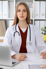  Doctor woman filling up medical form or typing on laptop while sitting at the table near the window. Medicine and health care concept