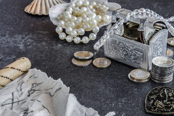 Fototapeta na wymiar Euro coins in the silver box and beside are a treasure with sea elements - shells on the dark table. At left side is a old treasure map written on the paper.