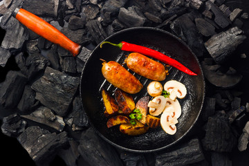 grilled sausages with potatoes, mushrooms and chili peppers on a grill pan with a background of charcoal
