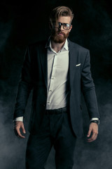Stylish handsome bearded man in trendy suit and eyeglasses posing in smoke