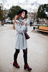 Young model tourist girl in a gray coat and black hat with leather handbag on shoulders posed at street of city.