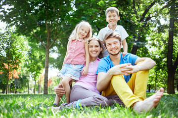 Fototapeta premium young happy family at noon in the park on the grass. Two young parents and children, boy and girl, sits on the grass and smiling looking at the camera.