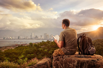 Fototapeta na wymiar hiker with backpack sitting on top of mountain enjoying view coast a modern city