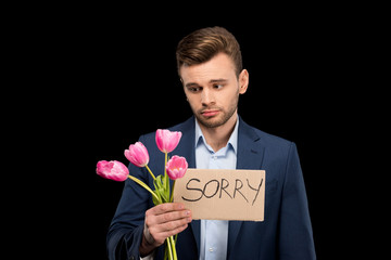 Upset young man holding pink tulips bouquet and sorry sign on black
