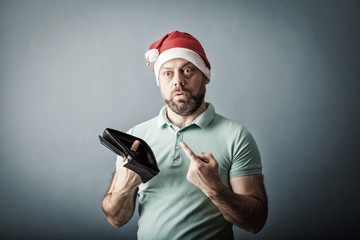 bearded middle-aged man in a santa hat sad, upset, unemployed broke guy, fired employee, holding looking into his empty wallet