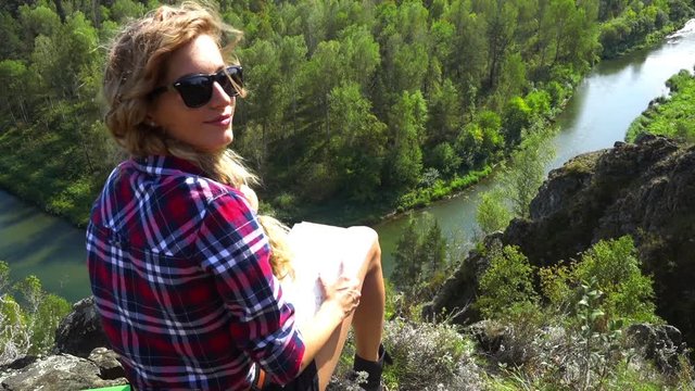 Young blonde woman tourist on a cliff over river Berd studying a map and view the neighborhood. Russia, Siberia, Salair