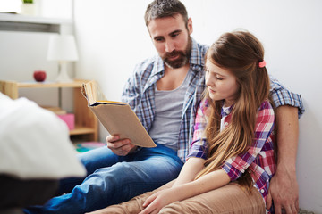 Handsome middle-aged father hugging his teenage daughter and reading exciting fairy-tale to her,...