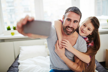 Beautiful little girl with long red hair hugging her joyful bearded dad and posing for selfie