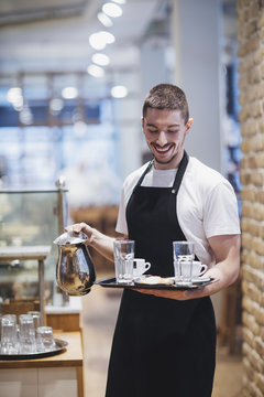 Smiling waiter holding tray and carafe at coffee shop