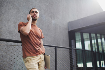Low angle view of bearded young man in casual clothing standing by metal fence with laptop and talking on phone
