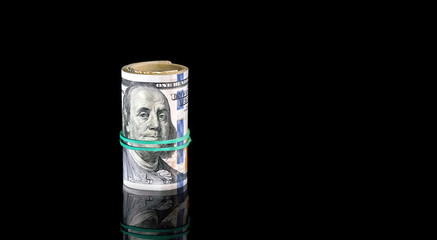 One hundred доллар bills rolled into a tube and tied by rubber band on the black reflective background.