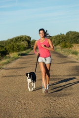 Naklejka na ściany i meble Sporty young woman running with her dog on a road. Female athlete training and exercising with a pet.