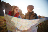Joyful travelers, young man and woman, taking a hike through mountains in bright sunlight, stopping to look at huge map with directions