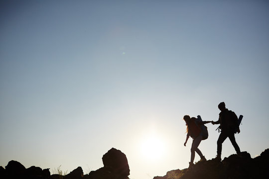 Backlit dark silhouettes of couple hiking in mountains, young man following girlfriends holding hands, side view graphic image