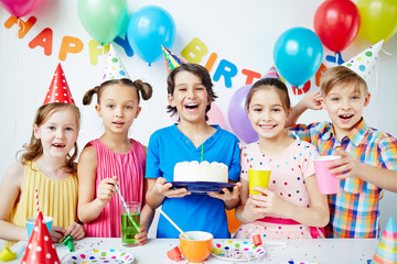 Group of agitated children together posing with birthday boy holding big birthday cake and looking at camera, all smiling cheerfully