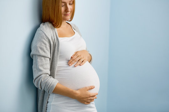 Gentle blond woman expecting baby, leaning on blue wall and looking at her big belly caringly