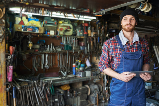Portrait of handsome bearded mechanic wearing uniform overalls standing holding digital tablet against background of shelves filled with tools in workshop