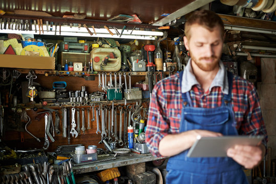 Modern bearded mechanic using tablet in workshop, focus on shelves with equipment and tools  behind him