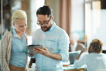 Waist-up portrait of male and female coworkers standing in meeting room and working on their business project with help of digital tablet