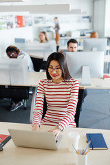 Pretty Asian woman in striped dress sitting at office desk and checking e-mails on laptop, her...