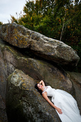 beautiful and young bride in white dress lying on big stone