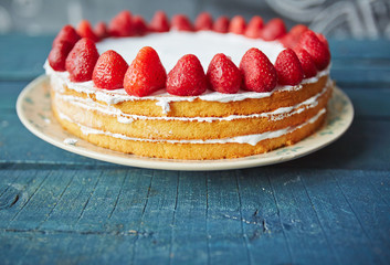 Closeup of delicious simple biscuit cake decorated with fresh ripe strawberries on top and white icing between layers standing on rustic wooden table