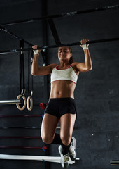 Fototapeta na wymiar Intense crossfit workout in dark gym: strong sportive woman performing pull-ups at wall mounted bar