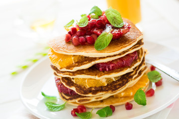 Traditional breakfast: stack of pancakes with orange slices and pomegranate seeds decorated mint leaves on white wooden table. Selective focus