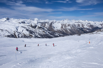 A wide piste, with a group of skiers on the piste, on a clear sunny day in Meribel, in the French Alps