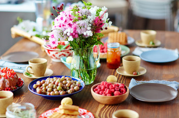 Rustic wooden table covered with bowls of freshly collected gooseberries and raspberries, bouquet of field flowers, homemade pies and cups of tea