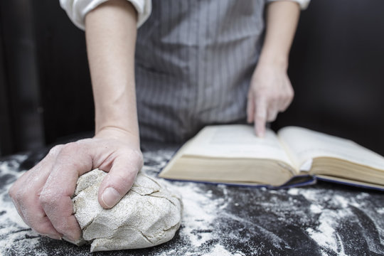 Baker chef looking for a recipe in a cookbook helping finger in the foreground is piece of dough on the black wooden table covered with flour