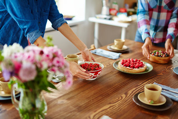 Women setting table with bowl of freshly collected redcurrant, delicious homemade tarts and cups of scented tea